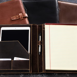 Pu&ograve; includere: Un portafoglio in pelle marrone con un blocco note integrato, un portapenne e una tasca per un tablet o un telefono. Il portafoglio ha una patta con una chiusura a fibbia.