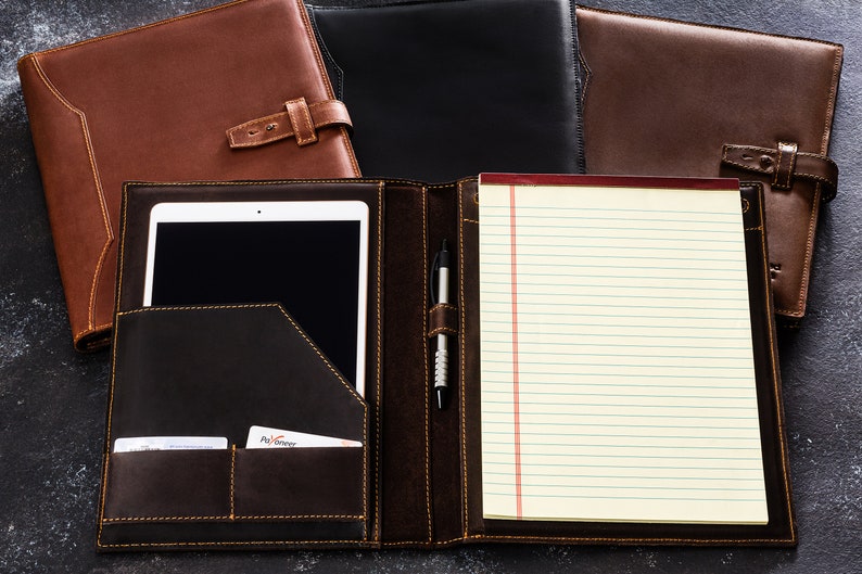 Pu&ograve; includere: Un portafoglio in pelle marrone con un blocco note integrato, un portapenne e una tasca per un tablet o un telefono. Il portafoglio ha una patta con una chiusura a fibbia.