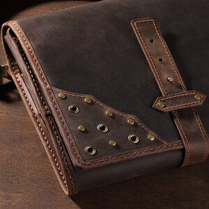 May include: A dark brown leather messenger bag with a shoulder strap. The bag features decorative brass rivets and stitching. A leather strap with a buckle secures the flap. The bag is designed with a vintage aesthetic.