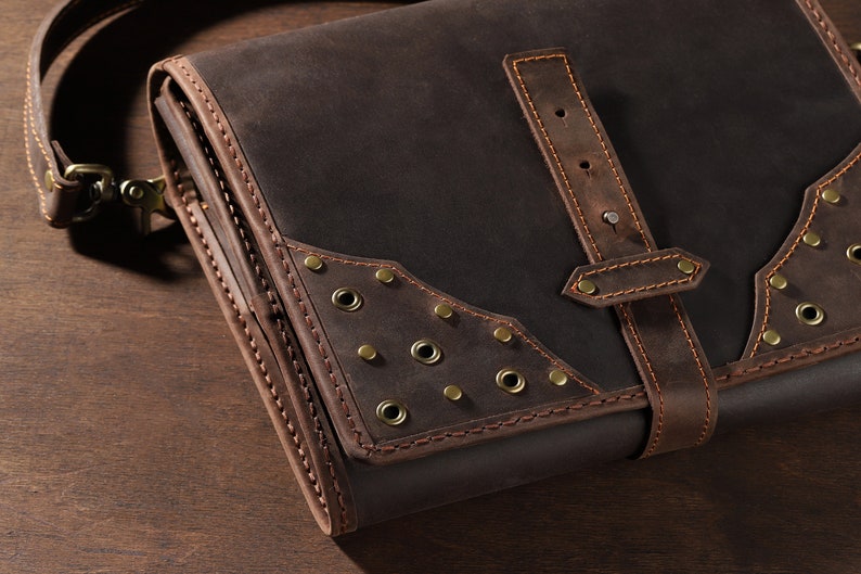 May include: A dark brown leather messenger bag with a shoulder strap. The bag features decorative brass rivets and stitching. A leather strap with a buckle secures the flap. The bag is designed with a vintage aesthetic.