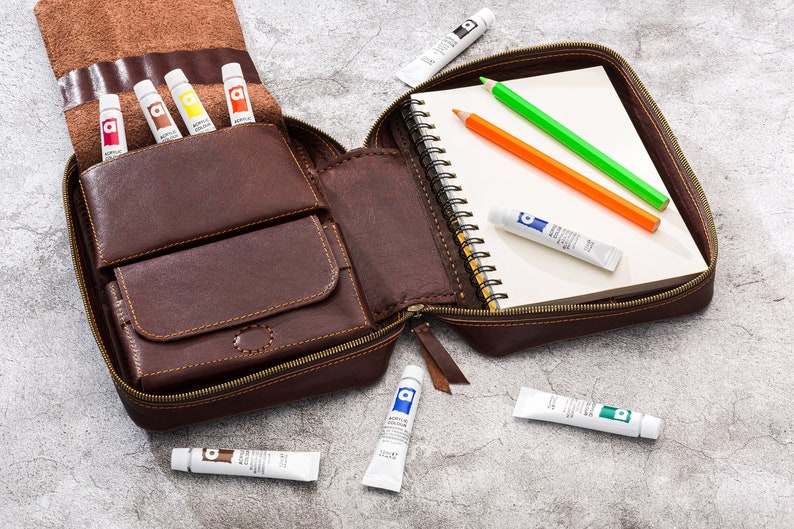 May include: A brown leather carrying case with a zipped opening. The case is open and contains a small sketchbook, coloured pencils, and tubes of acrylic paint.