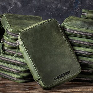 May include: A stack of green leather zippered pouches with the brand name "WOODBRINE" embossed on the front of the top pouch.