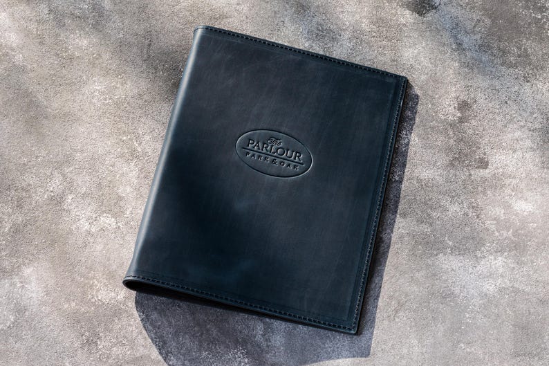 May include: A rectangular black leather menu cover featuring a debossed oval logo in the center with the text PARLOUR PARK & OAK. The cover has stitched edges and a smooth, matte finish, presenting a professional and clean appearance for document storage.