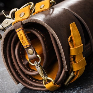 K&ouml;nnte beinhalten: Eine braune Lederrolle mit gelben Lederriemen und Schnallen. Die Rolle ist aufgerollt und mit einem Riemen gebunden.