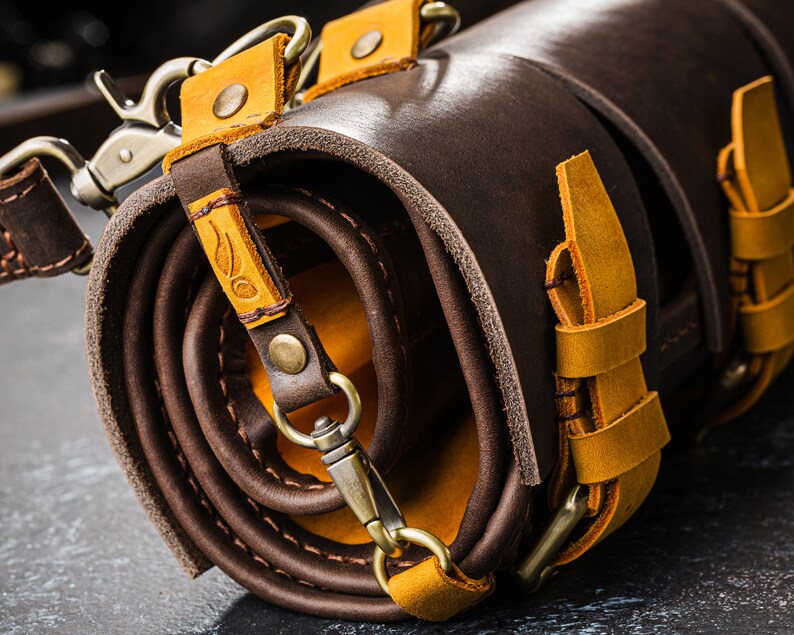 K&ouml;nnte beinhalten: Eine braune Lederrolle mit gelben Lederriemen und Schnallen. Die Rolle ist aufgerollt und mit einem Riemen gebunden.