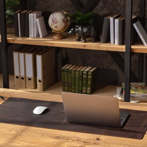 May include: A brown leather desk mat with a laptop computer and a white wireless mouse on a wooden desk. The desk is in front of a bookshelf with books, binders, and a globe.