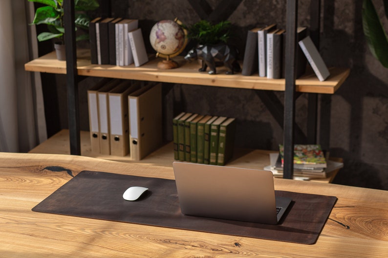 May include: A brown leather desk mat with a laptop computer and a white wireless mouse on a wooden desk. The desk is in front of a bookshelf with books, binders, and a globe.