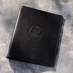 May include: A black leather menu cover or folder featuring a circular embossed logo in the center with the text THE BAY STREET. The rectangular item has stitched edges and a smooth, matte finish, presenting a professional and clean appearance.