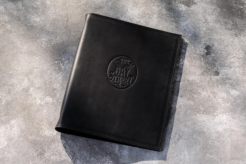 May include: A black leather menu cover or folder featuring a circular embossed logo in the center with the text THE BAY STREET. The rectangular item has stitched edges and a smooth, matte finish, presenting a professional and clean appearance.