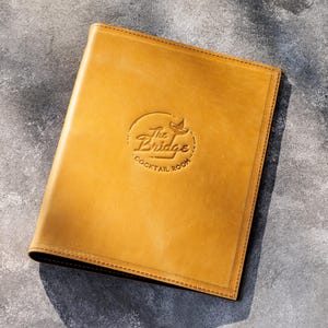 May include: A rectangular, mustard-yellow leather menu cover featuring a debossed circular logo in the center. The logo displays the text The Bridge Cocktail Room with a small martini glass icon. The cover has visible stitching along the edges.