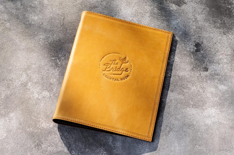May include: A rectangular, mustard-yellow leather menu cover featuring a debossed circular logo in the center. The logo displays the text The Bridge Cocktail Room with a small martini glass icon. The cover has visible stitching along the edges.