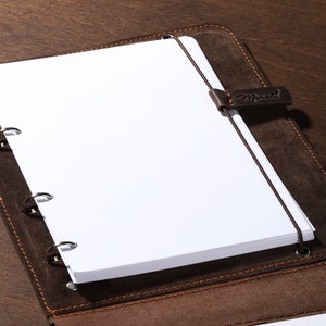 May include: A brown leather notebook with a stack of white paper and a green pencil. The notebook has a strap with the word "Manutti" on it. The notebook is open, revealing the blank pages and the pencil is resting on the side.