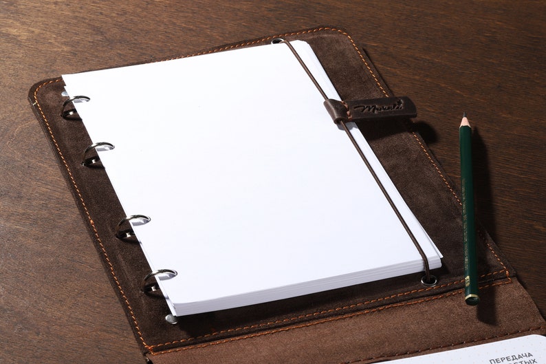 May include: A brown leather notebook with a stack of white paper and a green pencil. The notebook has a strap with the word "Manutti" on it. The notebook is open, revealing the blank pages and the pencil is resting on the side.