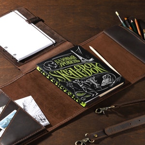 May include: A brown leather sketchbook with a notepad, pencil case, and pockets. The sketchbook has a black cover with green and white text. A pencil and several art supplies are visible.
