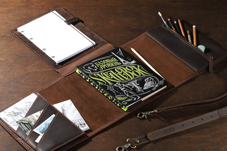 May include: A brown leather sketchbook with a notepad, pencil case, and pockets. The sketchbook has a black cover with green and white text. A pencil and several art supplies are visible.