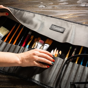 May include: A gray leather roll-up case with a black strap and the word "Leafy" embossed on the front. The case is open and filled with a variety of paint brushes.