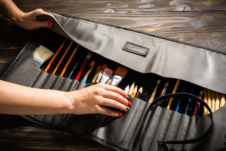 May include: A gray leather roll-up case with a black strap and the word "Leafy" embossed on the front. The case is open and filled with a variety of paint brushes.