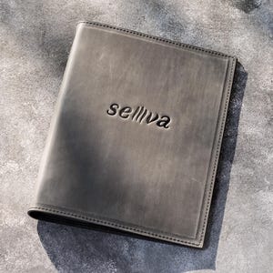 May include: A rectangular, dark gray leather folder featuring a smooth, matte finish and visible stitching along the edges. The word "selliva" is embossed in a lowercase, sans-serif font in the center of the front cover of the leather folder.