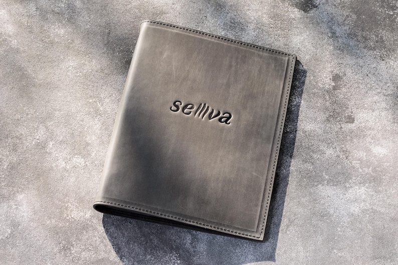 May include: A rectangular, dark gray leather folder featuring a smooth, matte finish and visible stitching along the edges. The word "selliva" is embossed in a lowercase, sans-serif font in the center of the front cover of the leather folder.
