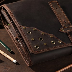 May include: A dark brown leather satchel with a flap closure and a shoulder strap. The satchel has decorative metal rivets and is surrounded by pencils and a pencil sharpener. The pencils are green, yellow, and brown.
