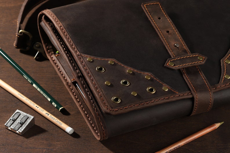 May include: A dark brown leather satchel with a flap closure and a shoulder strap. The satchel has decorative metal rivets and is surrounded by pencils and a pencil sharpener. The pencils are green, yellow, and brown.