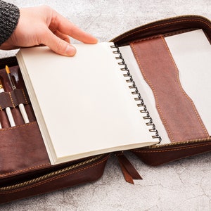 May include: A brown leather case with a zip fastening, containing a spiral bound notebook and a set of paintbrushes.