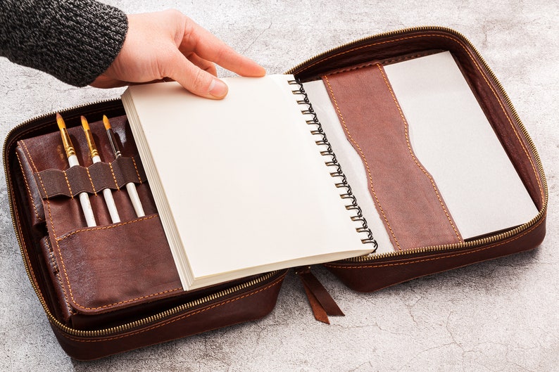 May include: A brown leather case with a zip fastening, containing a spiral bound notebook and a set of paintbrushes.