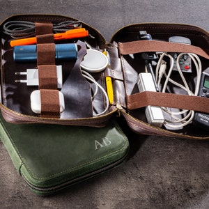 Op de afbeelding: Een bruine leren reistas met een ritssluiting en een groene leren etui met de initialen "AB" erop gegraveerd. De tas is open en gevuld met verschillende elektronische accessoires, waaronder snoeren, opladers en een klein apparaat.