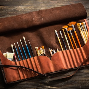 May include: A brown leather roll-up case with a strap closure, containing a variety of paintbrushes and pencils. The case is open and the brushes are visible.