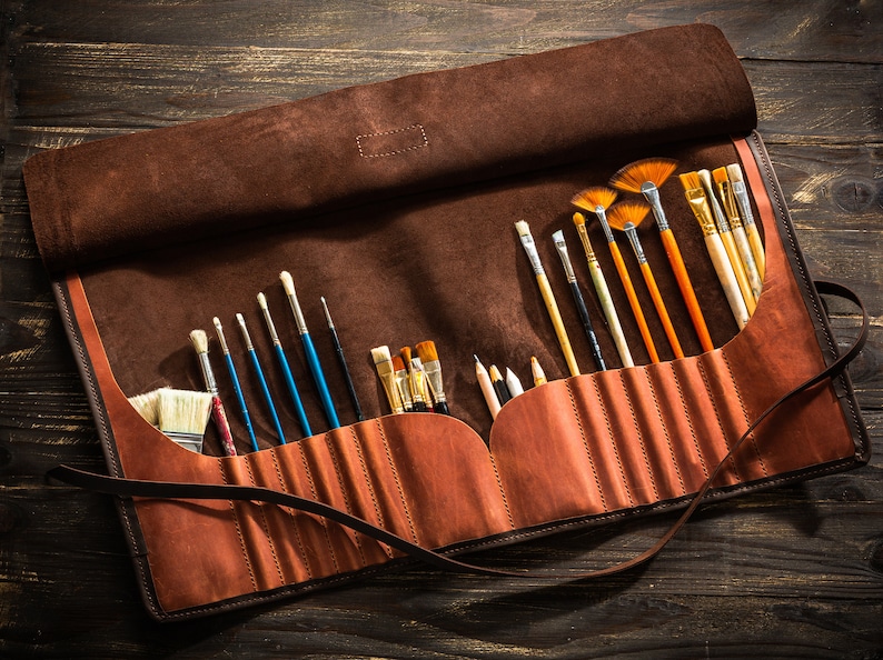 May include: A brown leather roll-up case with a strap closure, containing a variety of paintbrushes and pencils. The case is open and the brushes are visible.
