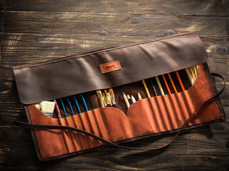 A brown leather brush roll-up case with a strap. The case is open and filled with paintbrushes and pencils. The case has a personalized name tag