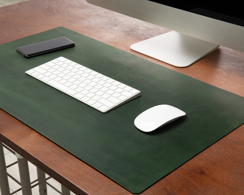 May include: A dark green desk mat with a white keyboard, mouse, and computer monitor. A black smartphone rests on the mat. The desk is made of wood.