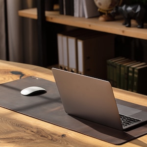 May include: A brown leather desk mat with a white computer mouse and a silver laptop on a wooden desk. The desk has a natural wood grain pattern. Shelves with books and office supplies are in the background.