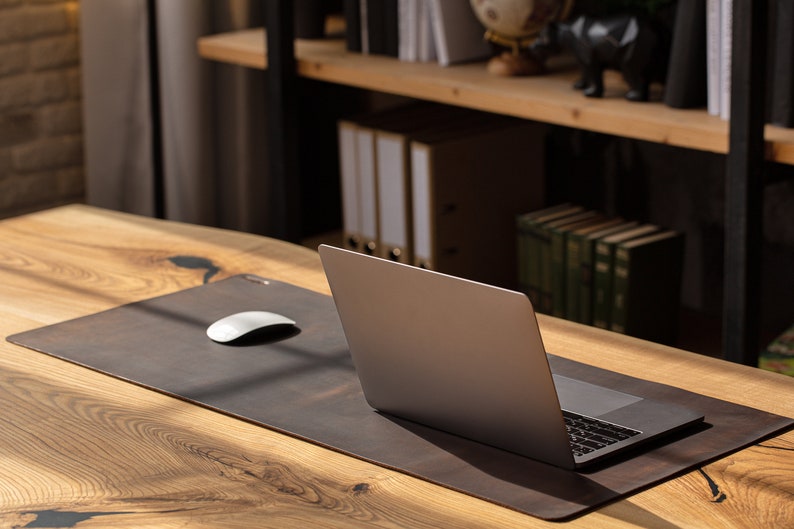 May include: A brown leather desk mat with a white computer mouse and a silver laptop on a wooden desk. The desk has a natural wood grain pattern. Shelves with books and office supplies are in the background.