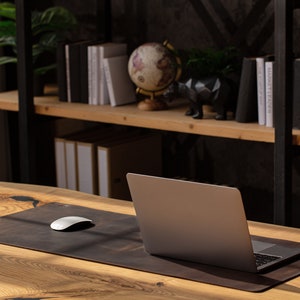 May include: A laptop sits on a brown leather desk mat with a white mouse. The desk is made of light-colored wood. A shelf in the background holds books, a globe, and decorative items, creating a workspace.