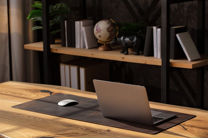 May include: A laptop sits on a brown leather desk mat with a white mouse. The desk is made of light-colored wood. A shelf in the background holds books, a globe, and decorative items, creating a workspace.