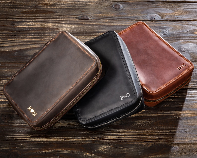 May include: Three leather zippered pouches with personalised initials. The pouches are brown, black, and dark brown. The initials are "ROM", "P&O", and "B+K".