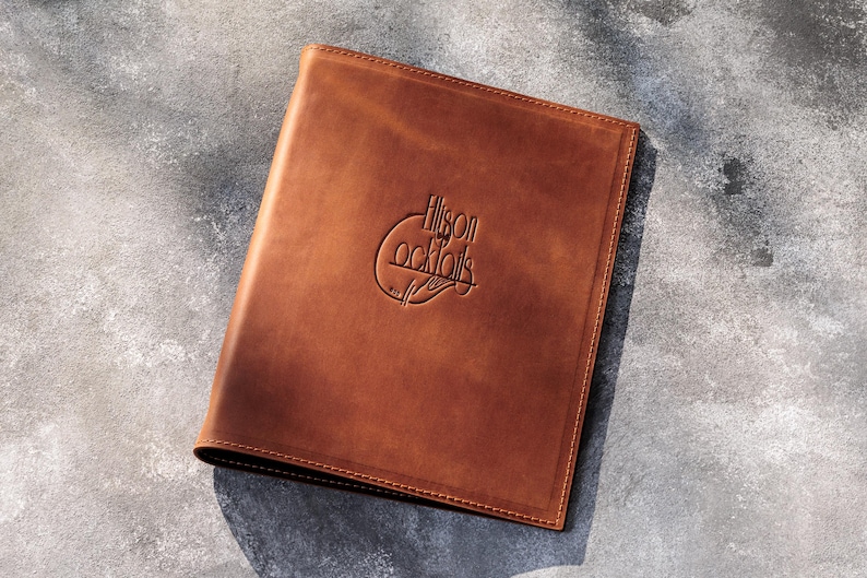 May include: A rectangular brown leather menu cover featuring a smooth, matte finish and stitched edges. The front cover is embossed with the text Ellison Cocktails, centered within a circular graphic design that includes a stylized hand illustration.
