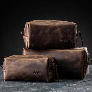 May include: Three brown leather toiletry bags stacked on top of each other. The bags have a rectangular shape with a zipper closure and a small handle. The top bag has the word "Vlabimir" embossed on it, and the bottom bag has the word "VOVA" embossed on it.