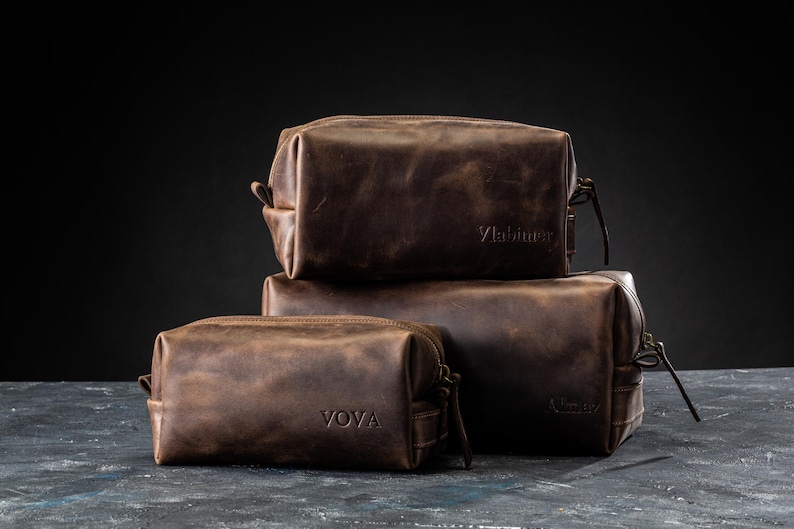 May include: Three brown leather toiletry bags stacked on top of each other. The bags have a rectangular shape with a zipper closure and a small handle. The top bag has the word "Vlabimir" embossed on it, and the bottom bag has the word "VOVA" embossed on it.