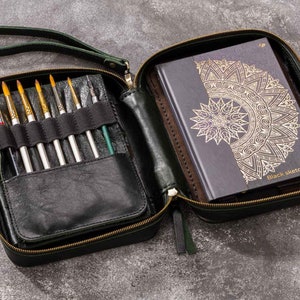 May include: A dark green leather zipped case with a black leather interior. The case is open and contains a set of paintbrushes and a black sketchbook with a gold mandala design. The sketchbook has the text "Black Sketch Note" on the cover.
