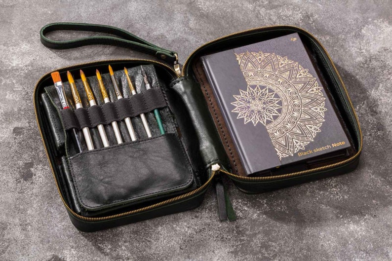 May include: A dark green leather zipped case with a black leather interior. The case is open and contains a set of paintbrushes and a black sketchbook with a gold mandala design. The sketchbook has the text "Black Sketch Note" on the cover.