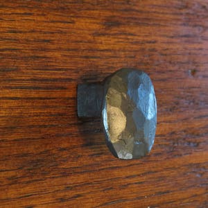 Op de afbeelding: Een gehamerde metalen kastknop met een ovale kop en een vierkante basis. De knop is donkergrijs en heeft een getextureerd oppervlak. De knop is gemonteerd op een houten oppervlak met zichtbare houtnerf.