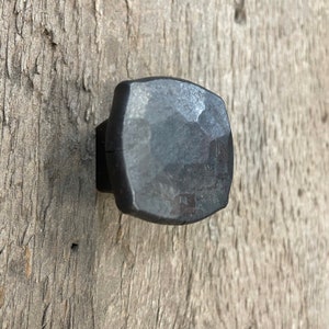 May include: A close-up shot of a dark grey, square metal cabinet knob with a hammered texture. It's mounted on a weathered, light brown wooden surface, giving a rustic appearance. The knob's design suggests a vintage or industrial style.