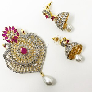 May include: A gold-tone peacock pendant necklace set with a white pearl drop and matching earrings. The pendant features a peacock design with pink and white stones. The earrings are also adorned with pink and white stones and have a white pearl drop.