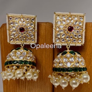 May include: Gold earrings with a square top and a round bottom. The square top is decorated with white stones and a pink stone in the center. The round bottom is decorated with white stones and a green border. The earrings are hanging from a string of white pearls.