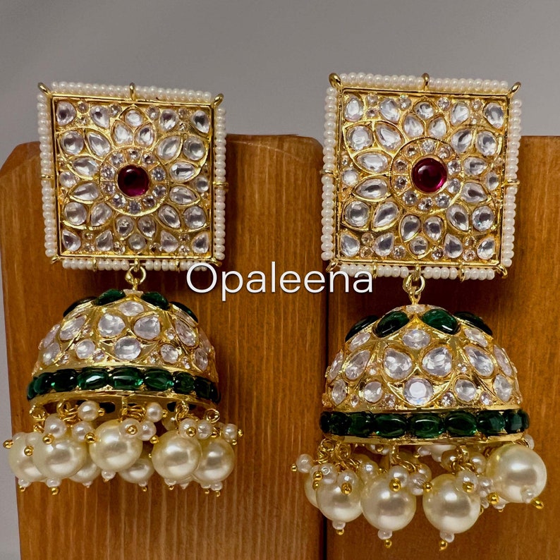 May include: Gold earrings with a square top and a round bottom. The square top is decorated with white stones and a pink stone in the center. The round bottom is decorated with white stones and a green border. The earrings are hanging from a string of white pearls.