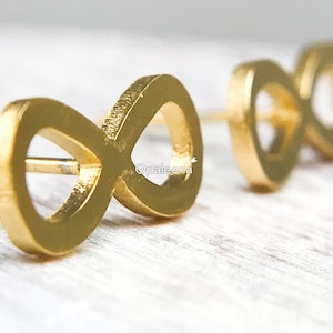 Könnte beinhalten: Ein Paar goldene Ohrstecker in Form eines Unendlichkeitszeichens. Die Ohrringe sind aus gebürstetem Metall und haben ein schlichtes, minimalistisches Design.