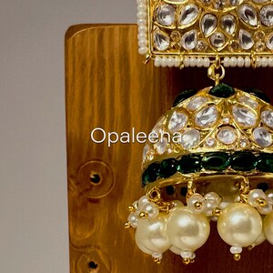 May include: Gold and green jhumka earrings with white pearl accents. The earrings feature a square top with a round bottom and are decorated with clear stones.