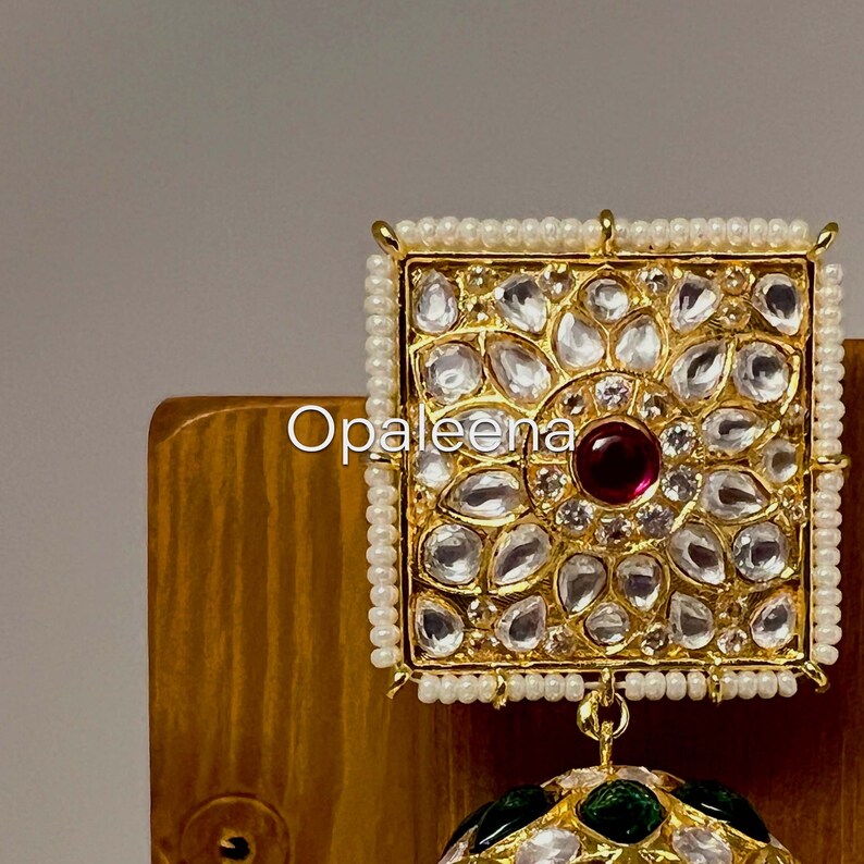 May include: Gold and white square earring with a red stone in the center and a round green and white earring hanging below. The square earring is surrounded by a border of white pearls.
